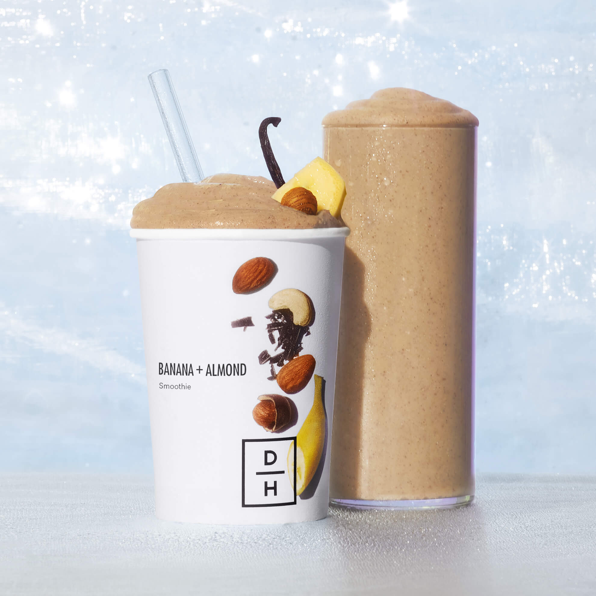 Daily Harvest Banana + Almond smoothie prepared in both a DH cup and a clear glass with blue and white sky background