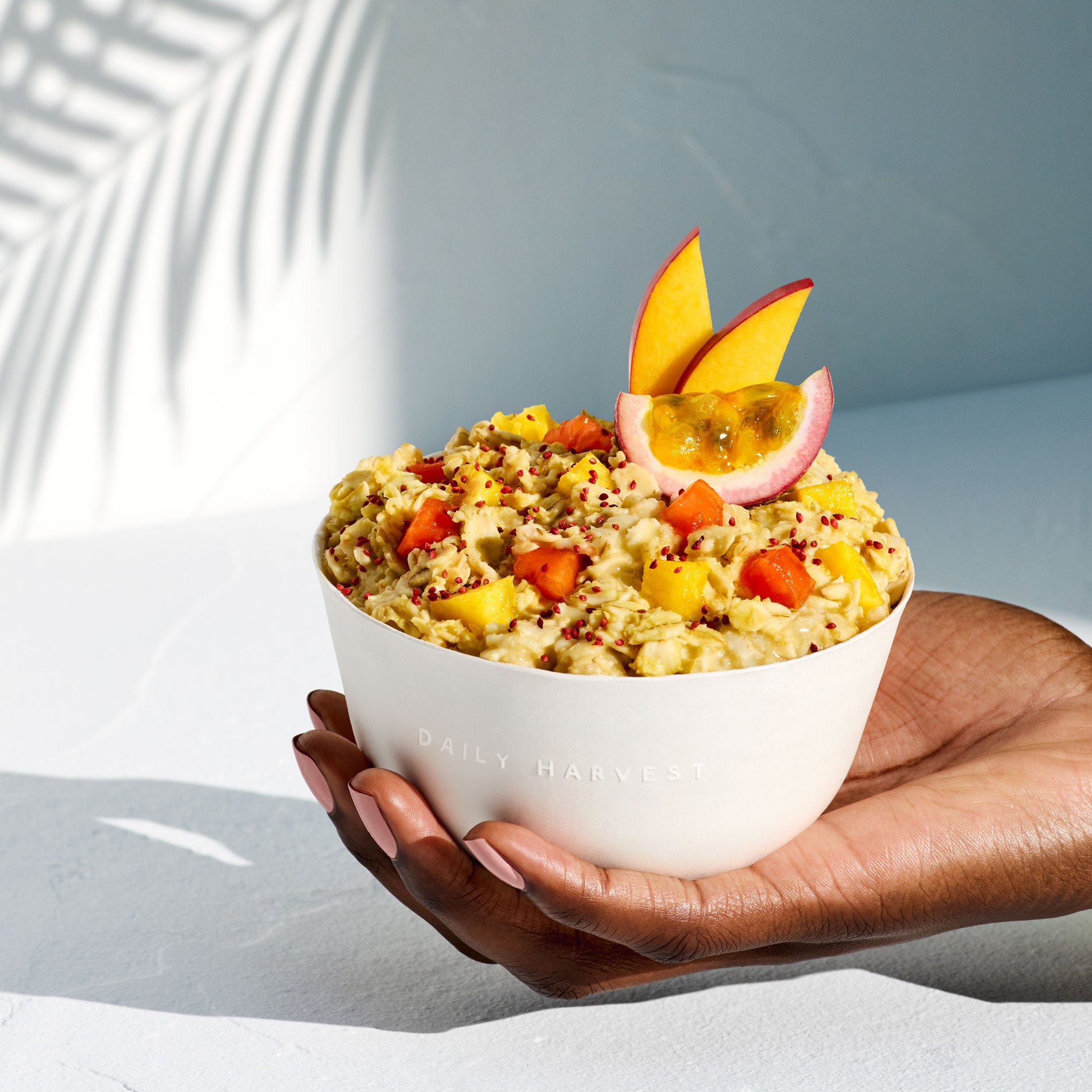 A hand holding a prepared Pineapple + Passion Fruit Forager Bowl in a white bowl with passion fruit and mango garnish.