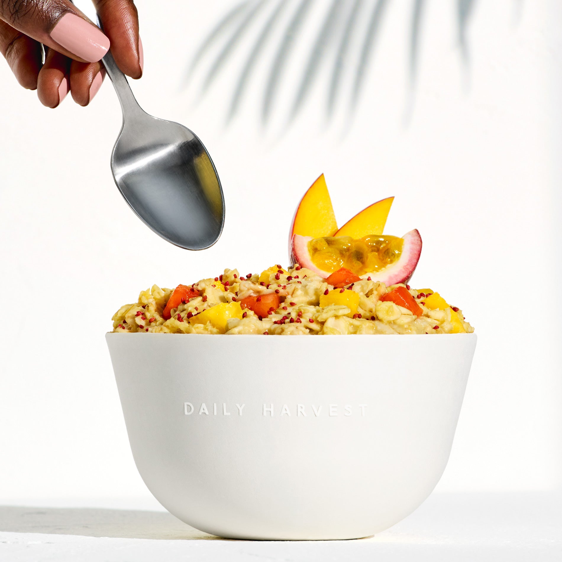 A hand with a spoon dipping into a prepared Pineapple + Passion Fruit Forager Bowl in a white bowl with passion fruit and mango garnish.