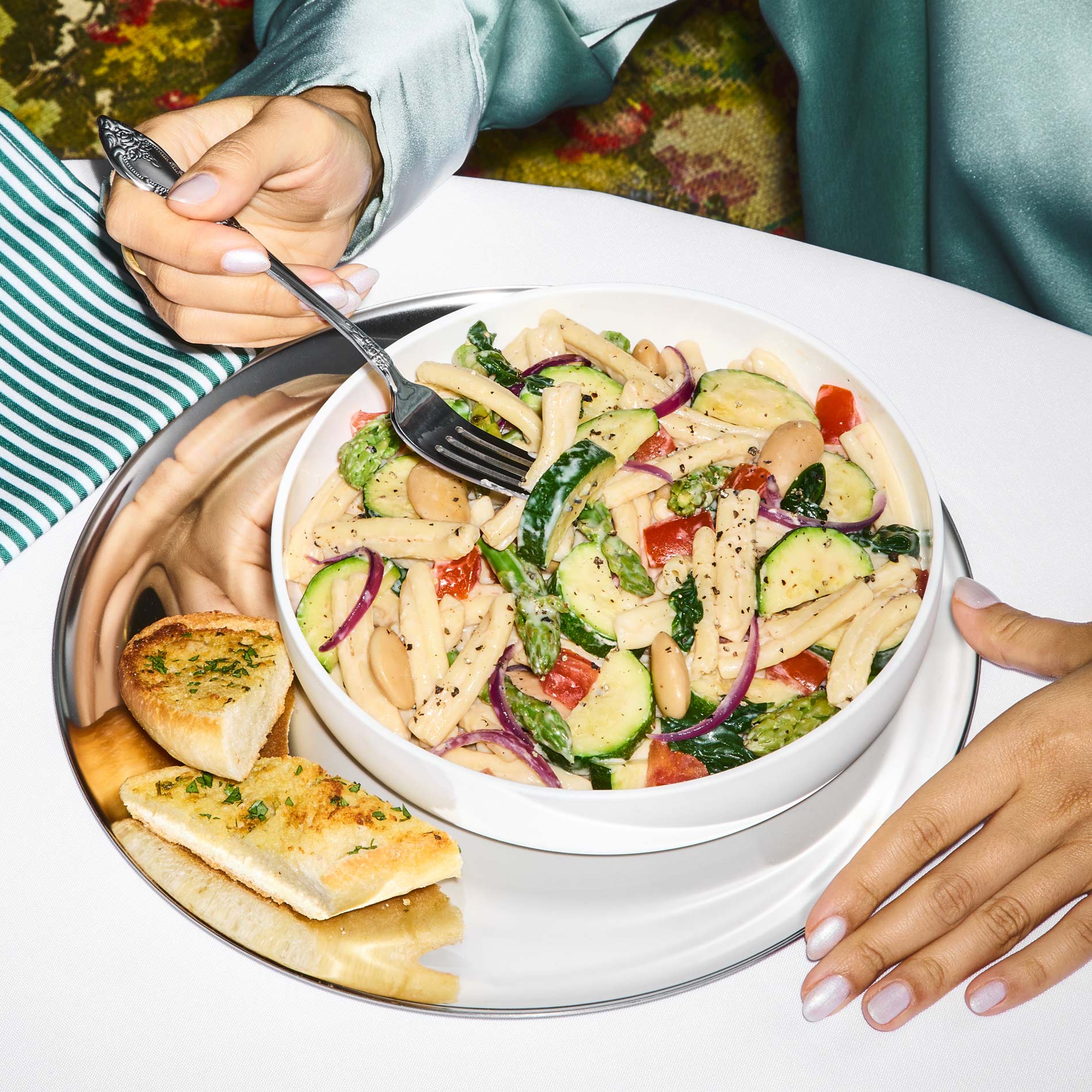 Hands holding a fork dipping into a prepared bowl of Zucchini + Gigante Bean Primavera Pasta with garlic bread on the side. 