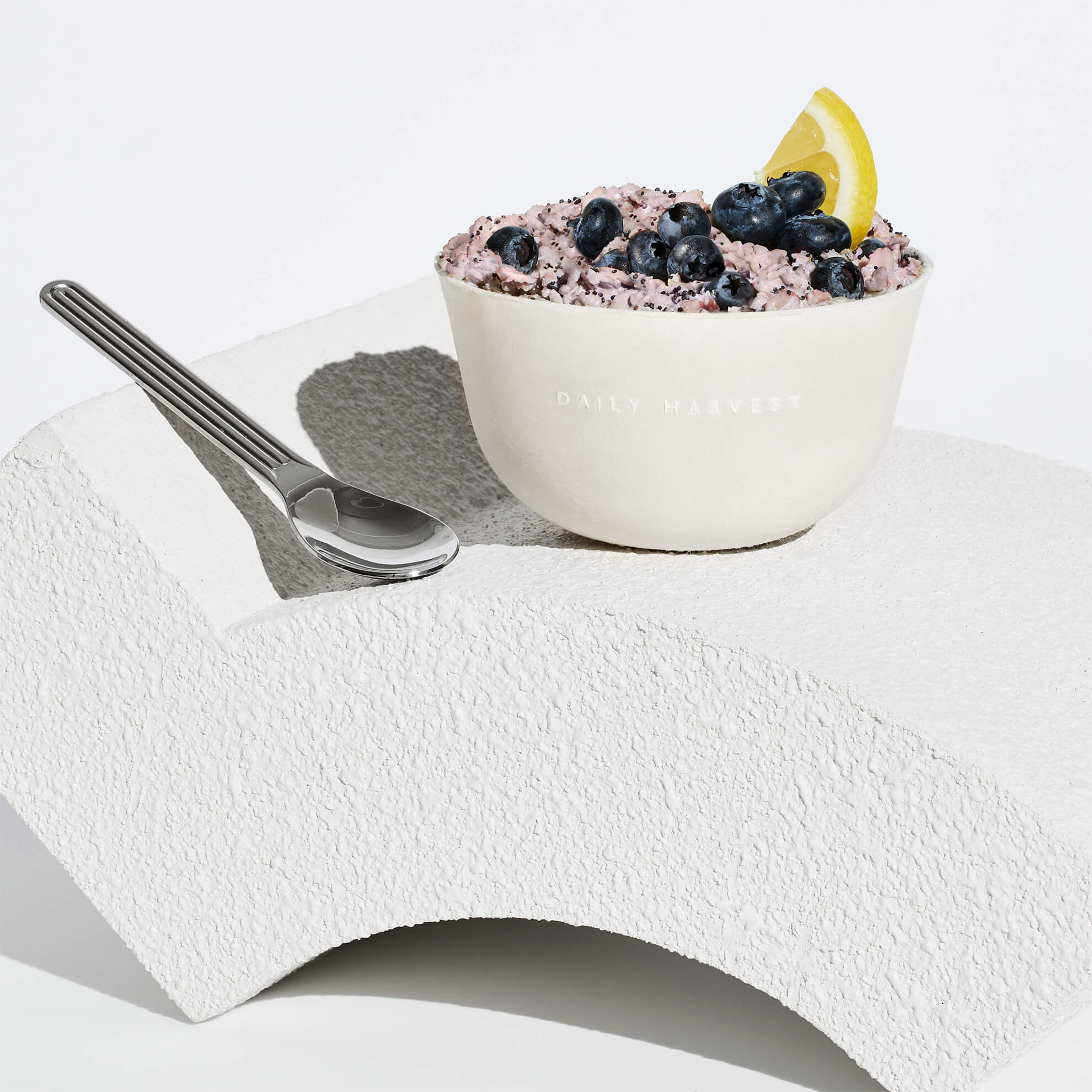 A lifestyle image of Daily Harvest Blueberry + Lemon Oat Bowl prepared with garnish on a white stand with a spoon.