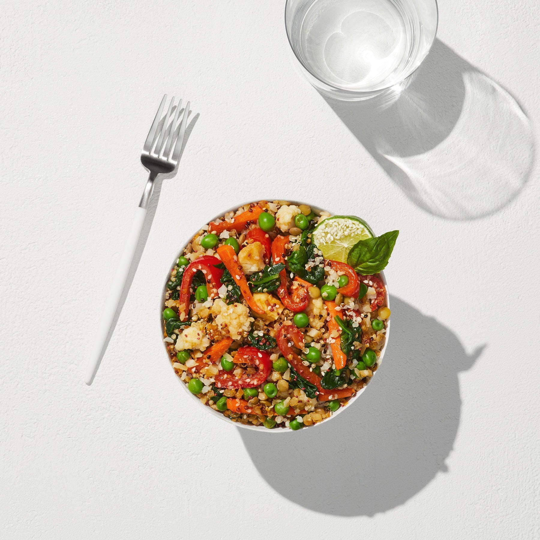 An overhead view of a fork, a glass of water, and a prepared Quinoa + Makrut Lime Harvest Bake in a large white bowl. 