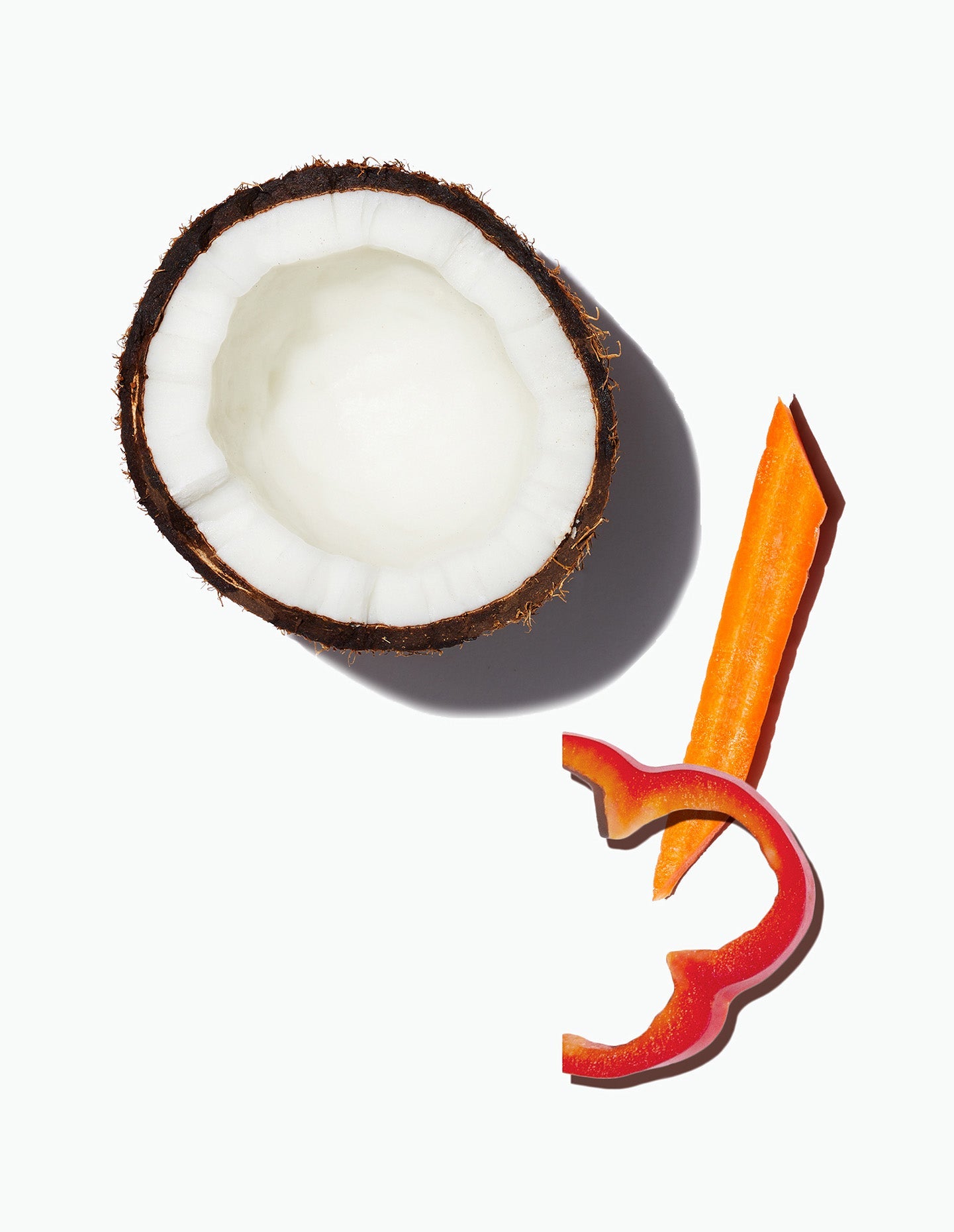 Image of a ingredients included in Daily Harvest Carrot + Coconut Curry soup including a  halved coconut with a slice of carrot and a small piece of red pepper. 