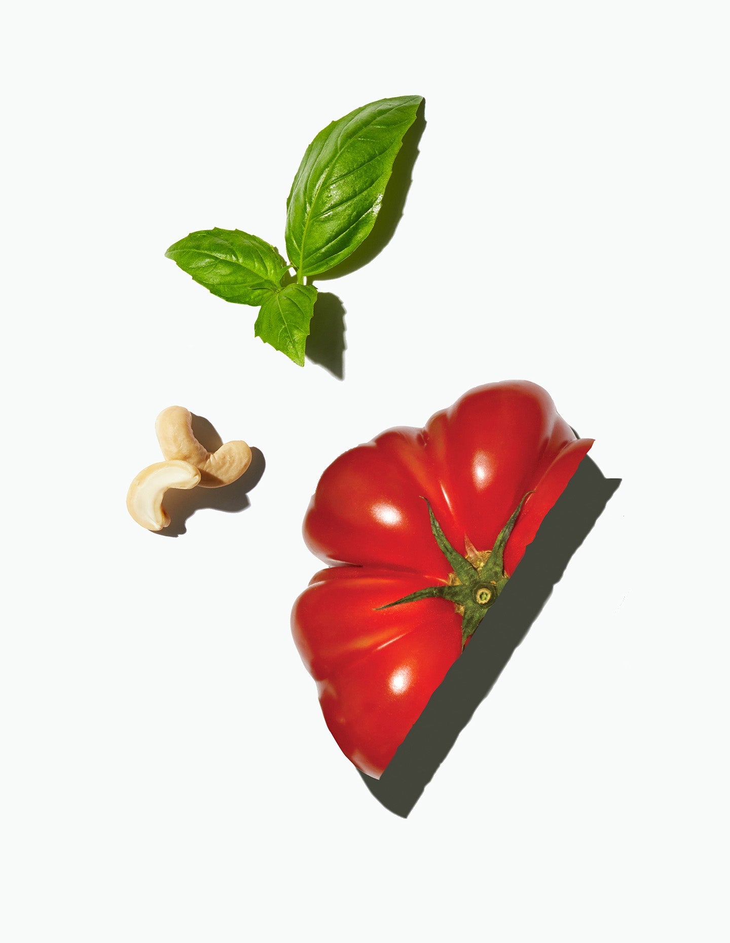 Image of Daily Harvest Cauliflower Rice + Pesto Harvest Bowl main ingredients, including basil leaves, a halved tomato, and a couple cashews. 