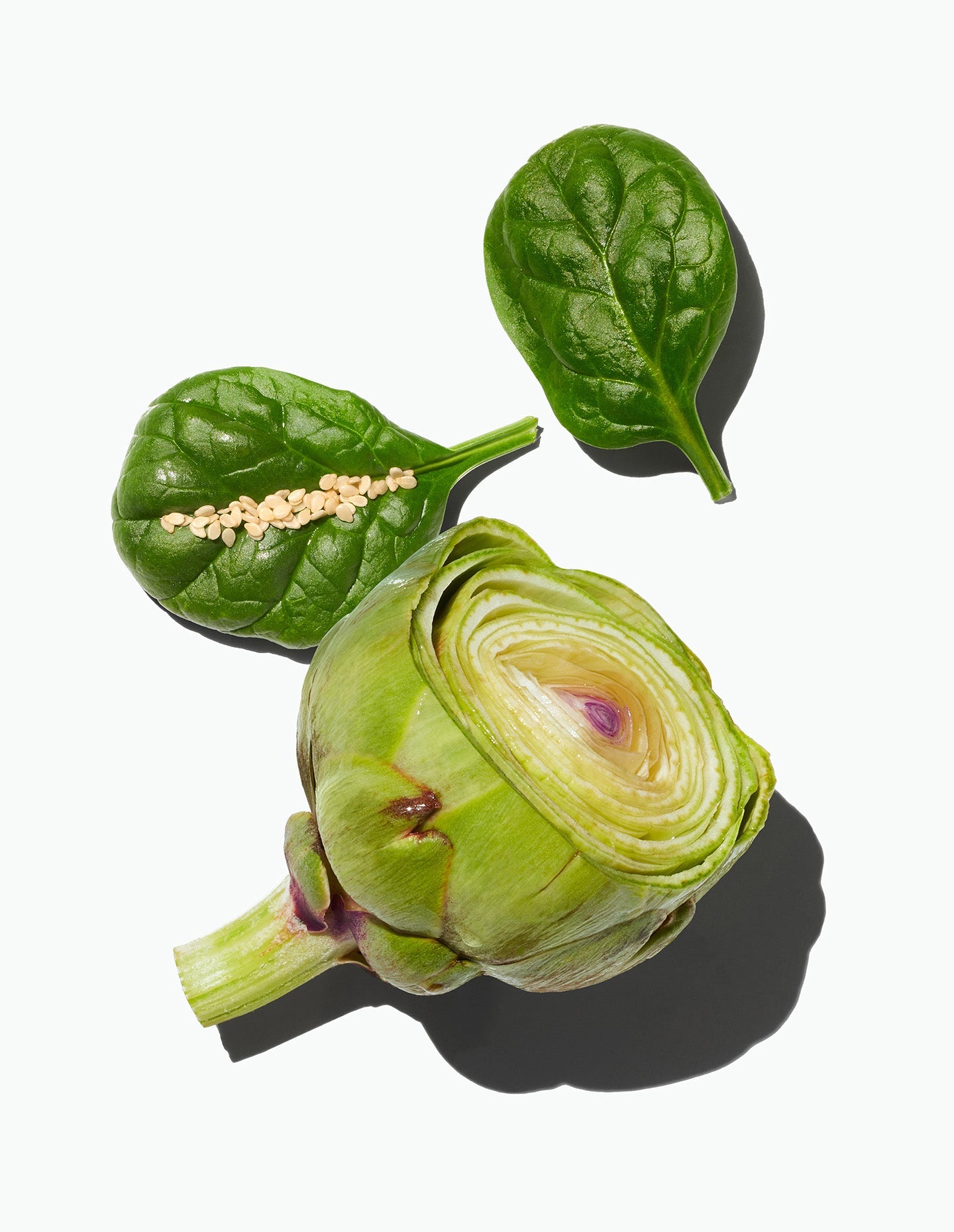 A sliced artichoke alongside spinach leaves and sesame seeds