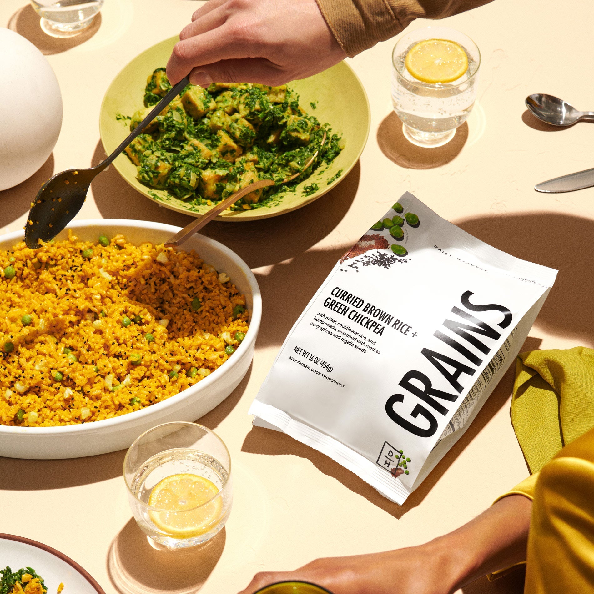 A picture of prepared Curried Brown Rice and Green Chickpea grains in a bowl on a table with water glasses and a plate of tofu next to the bag of grains.